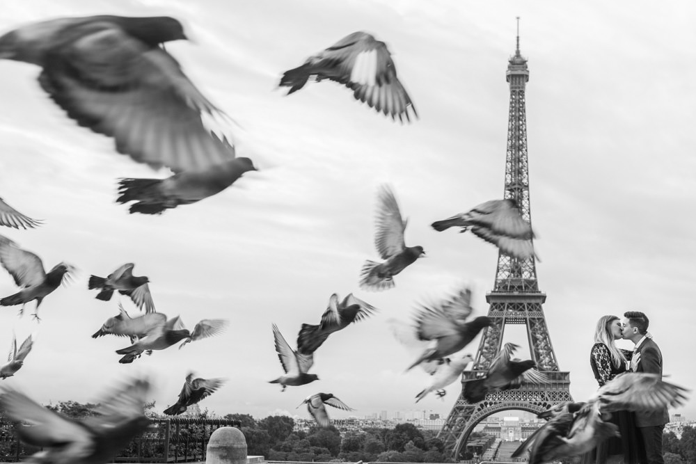 Couple kissing in front of the Eiffel Tower with pigeons flying around.